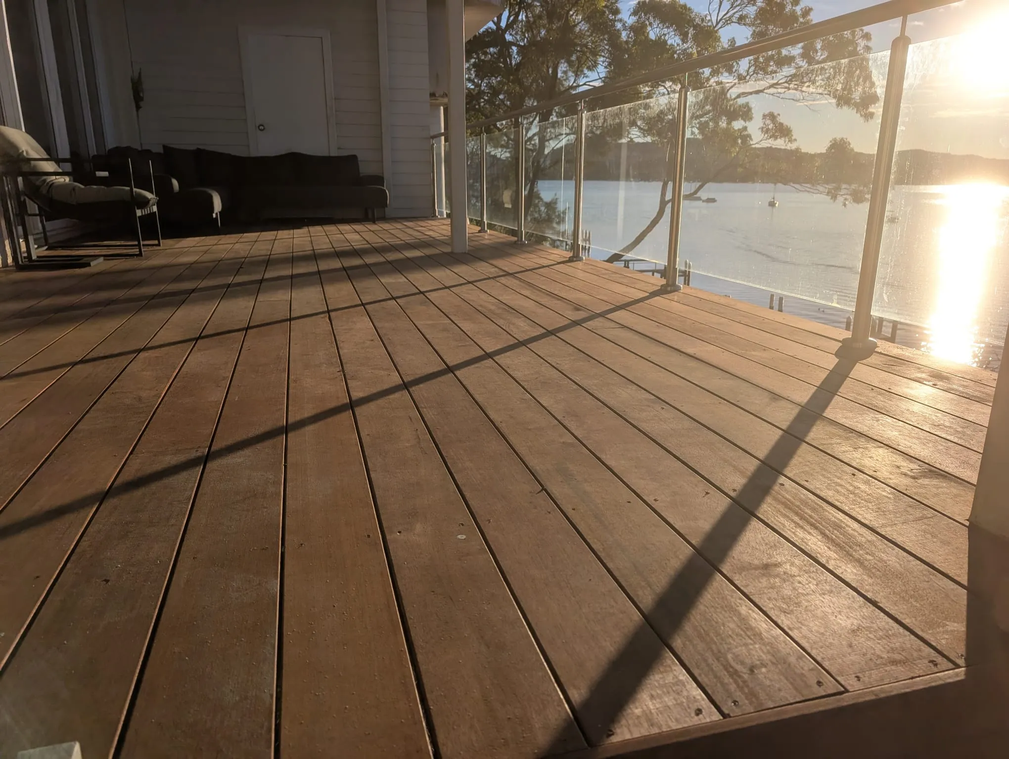 A renewed timber deck with a glass balustrade, refinished and lit by sunset over the water